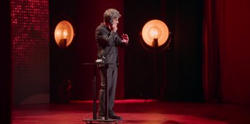 Movie still from “All About Gila” (2022), directed by Arturo González-Campos – A man standing on a stage holding a microphone; Wide shot, High angle