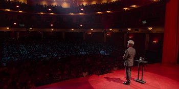 Movie still from “All About Gila” (2022), directed by Arturo González-Campos – A man standing on a stage in front of an audience; Extreme Wide shot, High angle