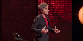 Movie still from “All About Gila” (2022), directed by Arturo González-Campos – A man standing on a stage wearing a black suit; Medium shot, High angle