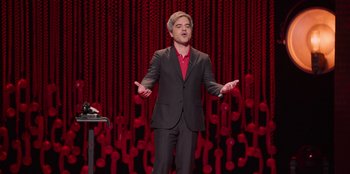 Movie still from “All About Gila” (2022), directed by Arturo González-Campos – A man standing in front of a microphone on a stage; Medium shot, High angle