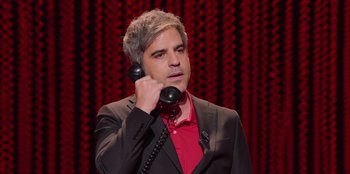 Movie still from “All About Gila” (2022), directed by Arturo González-Campos – A man holding a phone to his ear while standing in front of a red curtain; Close Up shot, High angle