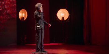 Movie still from “All About Gila” (2022), directed by Arturo González-Campos – A man standing on a stage holding a microphone; Wide shot, High angle