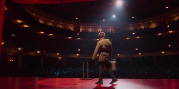 Movie still from “All About Gila” (2022), directed by Arturo González-Campos – A man standing on a stage in front of an audience; Wide shot, Low angle