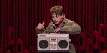 Movie still from “All About Gila” (2022), directed by Arturo González-Campos – A man in a brown jacket and a red neck tie is playing a radio; Medium shot, Over the shoulder angle