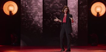 Movie still from “All About Gila” (2022), directed by Arturo González-Campos – A woman standing on a stage with her hands in the air; Wide shot, Over the shoulder angle