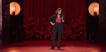 Movie still from “All About Gila” (2022), directed by Arturo González-Campos – A woman standing on a stage holding a cell phone to her ear; Wide shot, High angle