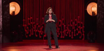 Movie still from “All About Gila” (2022), directed by Arturo González-Campos – A woman standing on a stage in front of a red background; Extreme Wide shot, High angle