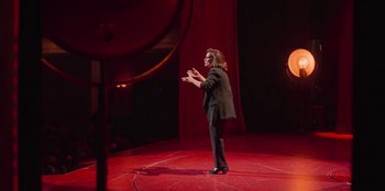 Movie still from “All About Gila” (2022), directed by Arturo González-Campos – A woman standing on a stage with her hands in the air; Wide shot, Over the shoulder angle
