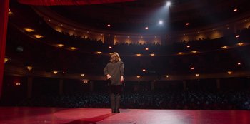 Movie still from “All About Gila” (2022), directed by Arturo González-Campos – A person standing on a stage in front of an audience; Extreme Wide shot, High angle