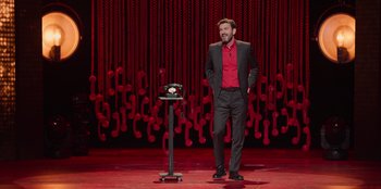 Movie still from “All About Gila” (2022), directed by Arturo González-Campos – A man standing on a stage next to an old fashioned phone; Wide shot, High angle