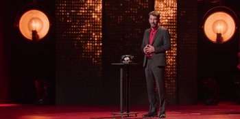 Movie still from “All About Gila” (2022), directed by Arturo González-Campos – A man standing on a stage in front of a microphone; Wide shot, High angle