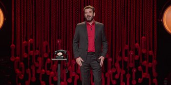Movie still from “All About Gila” (2022), directed by Arturo González-Campos – A man standing in front of a phone on a red background; Medium shot, High angle