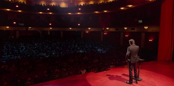 Movie still from “All About Gila” (2022), directed by Arturo González-Campos – A man standing on a stage in front of an audience; Extreme Wide shot, High angle