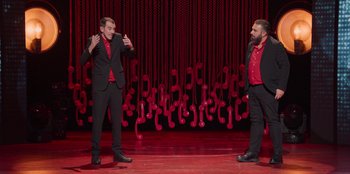 Movie still from “All About Gila” (2022), directed by Arturo González-Campos – A couple of men standing next to each other on a stage; Medium shot, Low angle