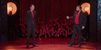 Movie still from “All About Gila” (2022), directed by Arturo González-Campos – Two men stand on a stage in front of a red background; Wide shot, High angle