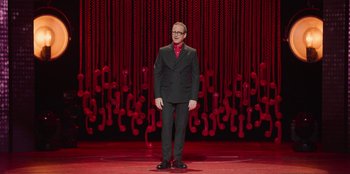 Movie still from “All About Gila” (2022), directed by Arturo González-Campos – A man standing on a stage wearing a suit and tie; Wide shot, High angle