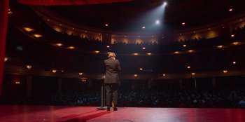 Movie still from “All About Gila” (2022), directed by Arturo González-Campos – A man standing on a stage in front of an audience; Wide shot, High angle