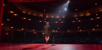 Movie still from “All About Gila” (2022), directed by Arturo González-Campos – A man standing on a stage holding a microphone; Extreme Wide shot, High angle
