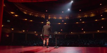Movie still from “All About Gila” (2022), directed by Arturo González-Campos – A man standing on a stage holding a microphone; Wide shot, High angle