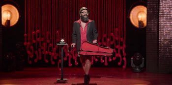 Movie still from “All About Gila” (2022), directed by Arturo González-Campos – A bearded man in a red suit carrying a guitar case; Wide shot, High angle