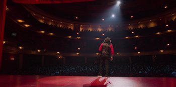 Movie still from “All About Gila” (2022), directed by Arturo González-Campos – A man standing on a stage in front of an audience; Extreme Wide shot, Low angle