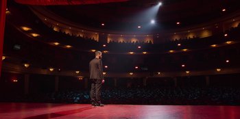 Movie still from “All About Gila” (2022), directed by Arturo González-Campos – A man standing on a stage in front of an audience; Extreme Wide shot, Low angle