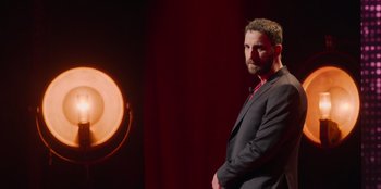Movie still from “All About Gila” (2022), directed by Arturo González-Campos – A man standing in front of an object on a stage; Medium shot, Over the shoulder angle
