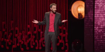 Movie still from “All About Gila” (2022), directed by Arturo González-Campos – A man standing on a stage with his arms outstretched; Medium shot, High angle