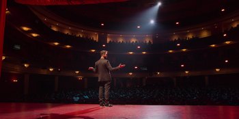 Movie still from “All About Gila” (2022), directed by Arturo González-Campos – A man standing on a stage in front of an audience; Extreme Wide shot, High angle