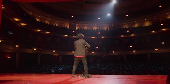 Movie still from “All About Gila” (2022), directed by Arturo González-Campos – A man standing on a stage with a microphone in front of a crowd; Extreme Wide shot, High angle