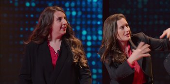 Movie still from “All About Gila” (2022), directed by Arturo González-Campos – Two women in black jackets and red shirts on a stage; Medium shot, Low angle