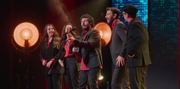 Movie still from “All About Gila” (2022), directed by Arturo González-Campos – A group of men and women singing on a stage; Wide shot, Low angle
