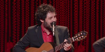 Movie still from “All About Gila” (2022), directed by Arturo González-Campos – A man in a red shirt is playing an acoustic guitar; Medium shot, Over the shoulder angle