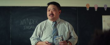 Movie still from “All the Bright Places” (2020), directed by Brett Haley – A man in a tie holding a book in front of a chalkboard; Close Up shot, Over the shoulder angle