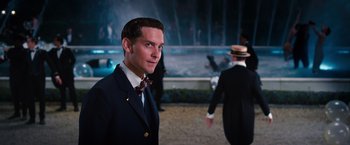 Movie still from “The Great Gatsby” (2013), directed by Baz Luhrmann – A man in a suit and tie standing in front of a fountain; Medium shot, Over the shoulder angle