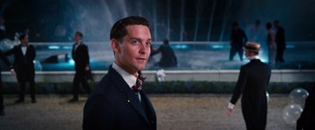 Movie still from “The Great Gatsby” (2013), directed by Baz Luhrmann – A man wearing a suit and tie standing in front of a fountain; Close Up shot, Over the shoulder angle