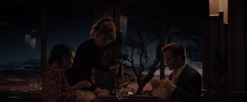 Movie still from “All the Old Knives” (2022), directed by Janus Metz – A woman standing at a table in front of two other people; Medium shot, Over the shoulder angle