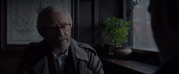 Movie still from “All the Old Knives” (2022), directed by Janus Metz – An older man wearing glasses sitting in front of a potted plant; Close Up shot, Over the shoulder angle
