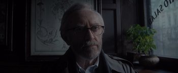 Movie still from “All the Old Knives” (2022), directed by Janus Metz – An old man with a beard wearing glasses and a jacket; Close Up shot, Over the shoulder angle