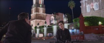 Movie still from “All the Places” (2023), directed by Pitipol Ybarra – A woman sitting on the back of a scooter in front of a building; Wide shot, Low angle