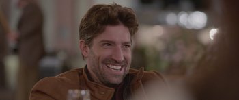 Movie still from “All the Places” (2023), directed by Pitipol Ybarra – A person wearing a brown jacket smiling; Close Up shot, Over the shoulder angle