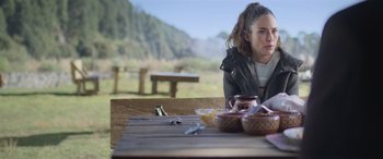 Movie still from “All the Places” (2023), directed by Pitipol Ybarra – A woman sitting at a wooden table with food on it; Medium shot, Over the shoulder angle