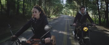 Movie still from “All the Places” (2023), directed by Pitipol Ybarra – Two people on motor scooters riding down the street; Wide shot, Low angle