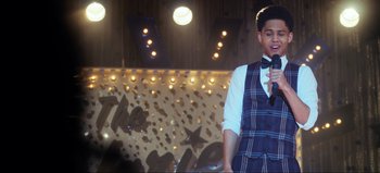 Movie still from “All Together Now” (2020), directed by Brett Haley – A man in a vest and bow tie is holding a microphone; Medium shot, Over the shoulder angle