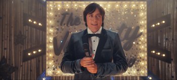 Movie still from “All Together Now” (2020), directed by Brett Haley – A man holding a microphone in front of a lighted wall; Medium shot, Over the shoulder angle