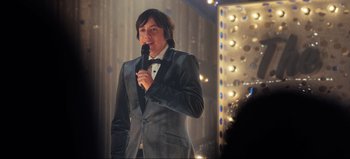 Movie still from “All Together Now” (2020), directed by Brett Haley – A man in a suit is holding a microphone; Medium shot, Over the shoulder angle
