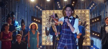 Movie still from “All Together Now” (2020), directed by Brett Haley – A young man in a plaid vest and bow tie holding a microphone; Medium shot, Over the shoulder angle