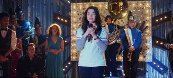 Movie still from “All Together Now” (2020), directed by Brett Haley – A woman holding a microphone in front of a group of people; Medium shot, Over the shoulder angle