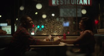 Movie still from “Along for the Ride” (2022), directed by Sofia Alvarez – Two people sitting at a table in front of a restaurant window; Medium shot, Over the shoulder angle