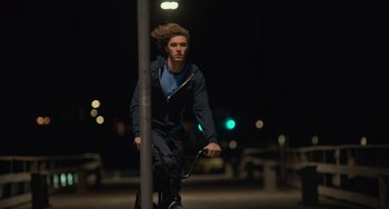 Movie still from “Along for the Ride” (2022), directed by Sofia Alvarez – A young man riding a bike down a street at night; Wide shot, Low angle